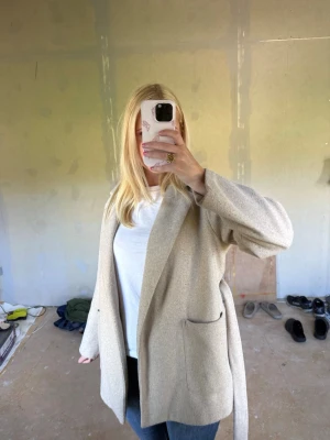 Beige fin kappa från ZARA med bälte - Stilren beige kappa från ZARA i storlek S. Kappan har en rak passform, stora fickor framtill och ett matchande bälte i midjan. Perfekt för lager-på-lager och enkel att styla till olika looks. Mjuk ullblandning som känns lyxig och bekväm. Hela jackan är tyvärr ganska nopprig därför det billiga priset