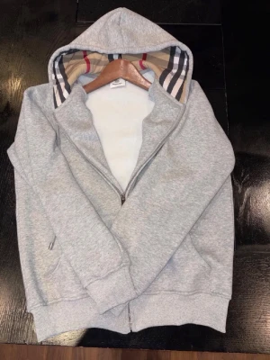 Går Burberry Zip Hoodie - Snygg grå zip hoodie från Burberry med klassiskt rutigt foder i huvan. Tillverkad i mjukt material med ribbade muddar och fickor på sidorna. Perfekt för dig som vill ha en stilren och exklusiv look med en twist av Burberrys ikoniska mönster.