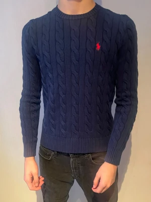 Mörkblå tröja Polo Ralph Lauren - Snygg mörkblå kabelstickad tröja från Polo Ralph Lauren med klassisk röd logga på bröstet. Tröjan har rund halsringning, långa ärmar och ribbade muddar. Perfekt att bära över en skjorta för en clean och stilren look.