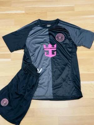 Svart och grått fotbollsställ - Säljer ett fotbollsställ med t-shirt och shorts i svart och grått. Tröjan har rosa logga och emblem på bröstet, samt korta ärmar. Shortsen har resårmidja och matchande emblem. Materialet är glansigt och andas, perfekt för träning eller match.