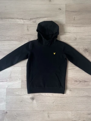 Svart hoodie från Lyle & Scott - Säljer en svart hoodie från Lyle & Scott med klassisk huva och känguruficka. På bröstet sitter den ikoniska gula örnloggan. Hoodien är i mjukt neoprene material och har ribbade muddar vid ärmar och nederkant. Perfekt för en chill och stilren look.
