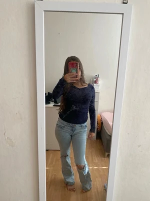 Ljusblå slitna bootcut jeans - Säljer ett par ljusblå bootcut jeans med slitningar och hål på ena knät. Jeansen har klassisk femficksdesign och är i en rak, lite utsvängd modell. Perfekta för en avslappnad och trendig look.