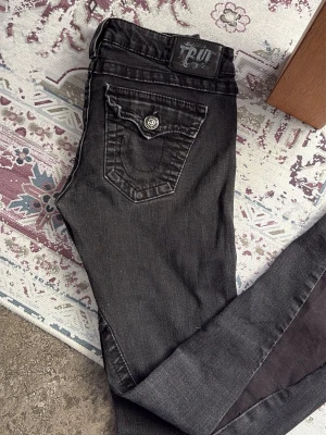 Svarta bootcut jeans True Religion - Säljer ett par svarta lågmidjade bootcut jeans från True Religion med ikonisk fickdesign och silverfärgad knappdetalj. Jeansen har låg midja och smal passform, perfekta till sneakers eller boots. Klassisk True Religion-lapp bak i midjan och snygga sömmar. Storlek 28, vet inte exakt vad det motsvarar