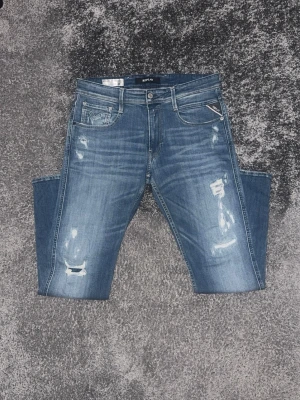 Blå slitna jeans från Replay - Säljer ett par blå jeans från Replay med slitningar och distressed detaljer på både fram- och baksida. Klassisk femficksmodell med normal midja och raka ben. Jeansen har en snygg tvättad look och Replay-lapp bak i midjan.