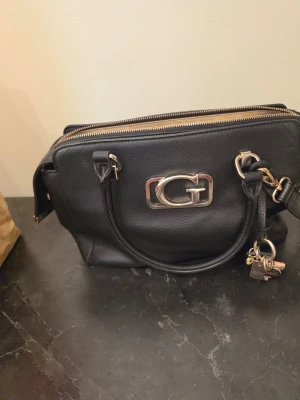 Äkta guess väska - Svart handväska i skinnimitation med guldfärgade metalldetaljer och ett stort G på framsidan. Väskan har både handtag och avtagbar axelrem samt dragkedja upptill. Dekorativ nyckelring med små berlocker hänger på sidan.