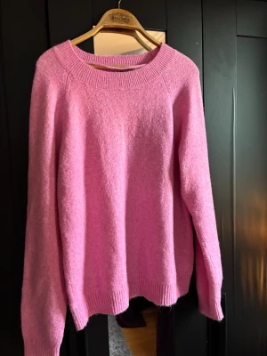 Rosa stickad tröja från Vero Moda - Mysig rosa stickad tröja från Vero Moda med rund halsringning och ribbade muddar vid ärmslut och nederkant. Tröjan har en avslappnad passform och är perfekt för att hålla värmen under kyliga dagar. Materialet är mjukt och behagligt.