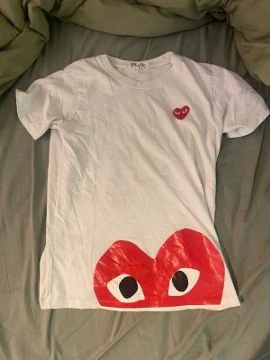 Cdg T-shirt  - Vit t-shirt med korta ärmar och ett stort rött hjärta med ögon tryckt längst ner samt ett mindre hjärta på bröstet. T-shirten har en klassisk passform och är gjord i mjuk bomull. Perfekt för dig som gillar grafiska tryck och vill sticka ut.