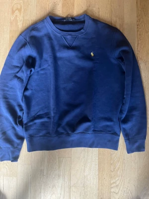 Mörkblå sweatshirt från Polo Ralph Lauren - Klassisk mörkblå sweatshirt från Polo Ralph Lauren med rund halsringning och den ikoniska broderade loggan i gult på bröstet. Tröjan har ribbade muddar vid ärmslut och nederkant. Perfekt för en avslappnad och stilren look.