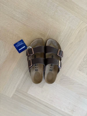 Bruna Birkenstock Arizona sandaler - Klassiska Birkenstock Arizona sandaler i mörkbrunt med två justerbara remmar och metallspännen. Ovandelen är i syntetiskt material och sulan har ett mönstrat grepp. Perfekta för en avslappnad och trendig look.