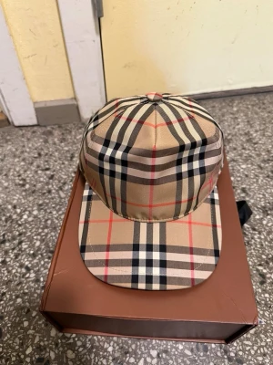 Burberry beige rutig keps - Snygg keps från Burberry i klassiskt beige rutigt mönster med svarta, vita och röda linjer. Justerbar baktill med svart plastspänne och Burberry London England-märke på baksidan. Perfekt accessoar för en trendig streetwear-look.
