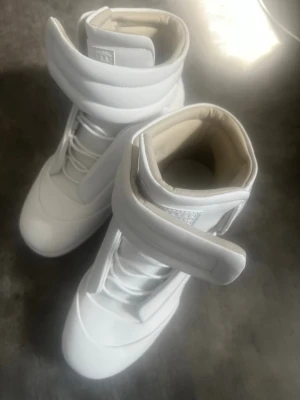 Vita höga sneakers maison Margiela - Säljer ett par helvita sneakers med högt skaft och breda kardborreband över vristen. Skorna har en futuristisk look med slät ovandel och tjock vaddering runt ankeln. Tillverkade i syntetmaterial och har vita snören. Perfekta för dig som gillar statement-skor med märket maison margiela.