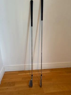 Två golfklubbor för 700kr tillsammans  - Säljer två golfklubbor med silverfärgade skaft i metall och svarta handtag. Klubborna har klassisk form med räfflade klubbhuvuden, perfekta för dig som vill börja spela golf eller behöver extra klubbor till din samling. K