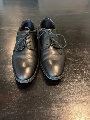 Svarta klassiska läderskor - Säljer ett par svarta klassiska läderskor med snörning och låg klack. Skorna har en stilren design med rundad tå och diskreta sömmar. Perfekta för dig som vill ha ett tidlöst och elegant par skor till garderoben.