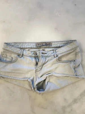Ljusblå denimshorts med rå kant - Säljer ett par ljusblå denimshorts med låg midja och rå, fransig kant. Klassisk femficksmodell med knapp och dragkedja framtill. Perfekta för varma dagar och enkel att matcha med allt.