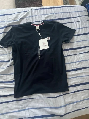 Svart Moncler t-shirt med logga - Svart t-shirt från Moncler med klassisk rund hals och liten Moncler-logga på bröstet. T-shirten har en diskret röd, vit och blå rand på insidan av kragen och är tillverkad i mjuk bomull. Perfekt för en clean och stilren look.
