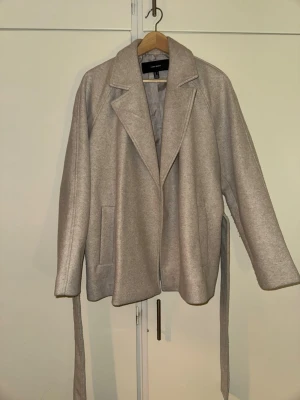 Beige kappa med knytskärp - Stilren beige kappa med bred krage och knytskärp i midjan. Kappan har raka ärmar, två sidofickor och en mjuk, något borstad yta. Perfekt för dig som vill ha en enkel men trendig look till hösten eller våren.