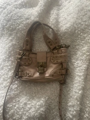 Beige handväska med nitar och spänne - Säljer en beige handväska i skinnimitation med coola nitar och detaljerade sömmar. Väskan har både handtag och en lång axelrem, samt ett dekorativt spänne framtill. Perfekt för dig som gillar edgy detaljer och praktisk design.