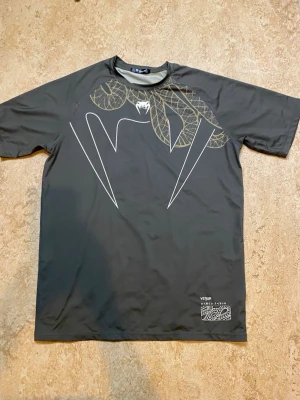 Svart Venum t-shirt med ormtryck - Cool svart t-shirt från Venum med stort grafiskt ormtryck både fram och bak. På ryggen står det 'VENUM' i stora bokstäver och en stor orm slingrar sig över hela tröjan. Tillverkad i mjukt syntetmaterial och har en avslappnad passform med korta ärmar.