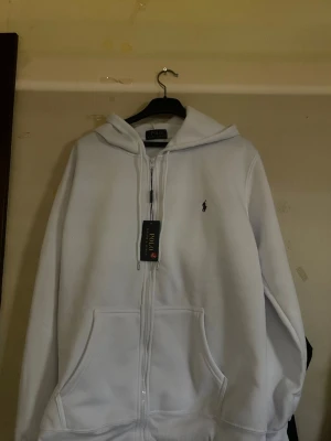 Vit hoodie från Polo Ralph Lauren - Snygg vit hoodie med dragkedja från Polo Ralph Lauren. Klassisk design med huva, två stora fickor framtill och den ikoniska lilla loggan broderad på bröstet. Tillverkad i mjukt material som känns skönt mot huden. Perfekt för en clean och stilren look.