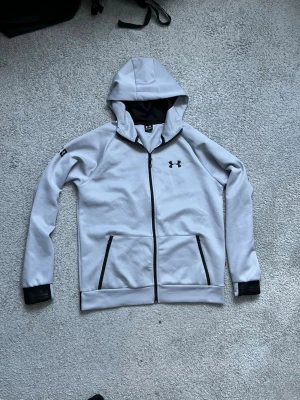 Ljusgrå hoodie från Under Armour - Snygg ljusgrå hoodie från Under Armour med dragkedja framtill och svarta detaljer. Två fickor med dragkedja, svart logga på bröstet och svart kant vid ärmslut. Huva och svart insida i huvan. Under Armour-tryck på ryggen längst ner. passar som xl/L
