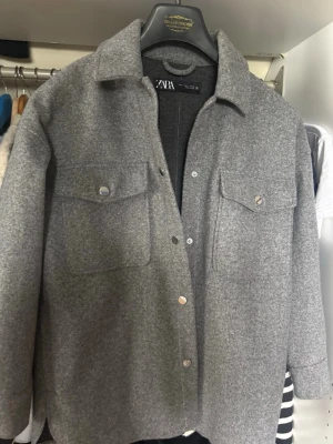 Grå overshirt/jacka från ZARA - Stilren grå overshirt från ZARA med klassisk krage och tryckknappar framtill. Två stora bröstfickor med lock och knappdetaljer. Overshirten har en rak passform och är gjord i ett mjukt, filtliknande material som ger en modern look.