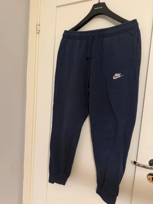Mörkblå Nike mjukisbyxor M - Säljer ett par mörkblå mjukisbyxor från Nike med dragsko i midjan och resår vid bensluten. Byxorna har två sidofickor och en broderad vit Nike-logga på vänster ben. Perfekta för chill dagar eller träning.