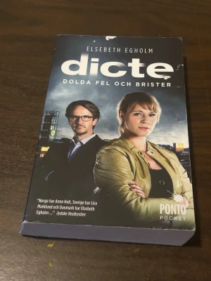 Dicte: Dolda fel och brister pocket - Spännande pocketbok av Elsebeth Egholm, 'Dicte: Dolda fel och brister'. Omslaget är mörkt med två personer i fokus och stadsmiljö i bakgrunden. Boken är på svenska och ges ut av Ponto Pocket. Perfekt för dig som gillar kriminalromaner och nordisk spänning.