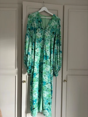 Grönblommig långklänning från Adoore - Långklänning från adoore i gröna och blå toner med blommigt mönster. Klänningen har v-ringning och långa, puffiga ärmar. Perfekt för sommarens alla tillfällen. Tillverkad i ett lätt och luftigt material.