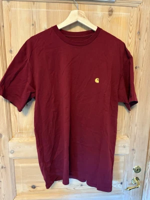 Vinröd Carhartt T-shirt - Säljer en vinröd T-shirt från Carhartt med klassisk rund hals och liten gul logga på bröstet. T-shirten är i mjuk bomull och har en enkel, clean design med normal passform och korta ärmar. Perfekt till jeans eller shorts.