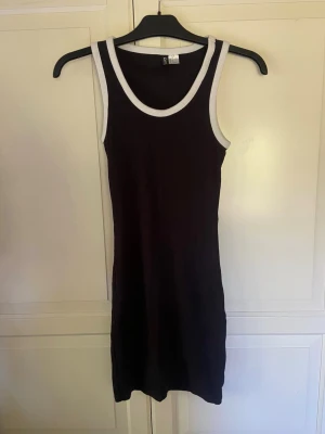 Svart ärmlös ribbad klänning - Snygg svart kortklänning med vita ribbade kanter vid hals och ärmhål. Klänningen är ärmlös och har en kroppsnära passform som ger en sportig och stilren look. Perfekt för varma dagar eller att styla med en jacka.