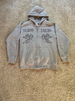 Grå rinestone Hoodie från bling lucid 140 - Säljer en grå zip-hoodie med huva och fickor framtill. På bröstet står det 'BLING' och 'LUCID' med två cupido-figurer i svart tryck. Hoodien har en avslappnad passform och är tillverkad i mjukt sweatshirtmaterial. Perfekt för en chill och trendig look.