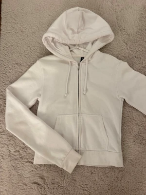 Vit zip hoodie  - Säljer en stilren vit hoodie med dragkedja framtill, två magfickor och justerbar huva med snören. Hoodien har långa ärmar och ribbade muddar vid ärmslut och nederkant. Perfekt för en avslappnad och clean look.