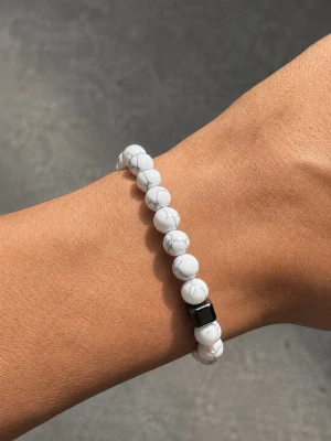 Vitt pärlarmband med marmormönster - Snyggt armband med vita runda pärlor som har ett marmorerat mönster och en svart fyrkantig pärla som detalj. Armbandet är elastiskt och passar de flesta handleder. Perfekt accessoar för att lyfta din stil med en clean och modern vibe.