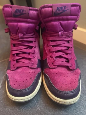 Nike dunk skor - Nike sneakers i high-modell med en mix av vinrött och lila. Ovandelen är i mocka och läder, med klassisk Nike Swoosh på sidan. Skorna har matchande vinröda skosnören och en vit sula med svart yttersula. Perfekta för dig som vill sticka ut med färg. De är en 2013 modell och svåra att får tag på
