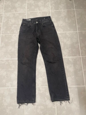 Svarta raka jeans från Zara - Säljer ett par svarta raka jeans från Zara i storlek 38. Jeansen har hög midja, klassisk femficksdesign och råa, fransiga benslut för en trendig look. Tillverkade i kraftigt denimtyg som ger en snygg vintagekänsla.