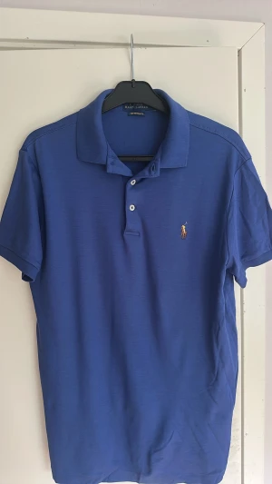 Blå pikétröja från Polo Ralph Lauren - Säljer en klassisk blå pikétröja från Polo Ralph Lauren med korta ärmar och krage. Tröjan har två knappar framtill och den ikoniska broderade loggan på bröstet. Tillverkad i mjukt supima cotton som är väldigt behalig att bära, en lite finare kvalité en deras vanliga. Knappt använd!
