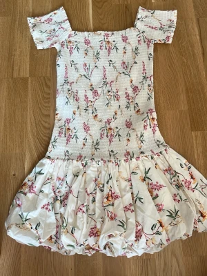 Blommig kortklänning med volang - Supersöt kortklänning med blommigt mönster i rosa, grönt och gult på vit botten. Klänningen har puffiga korta ärmar, smockad överdel och volangig kjol. Perfekt för sommarens alla tillfällen!