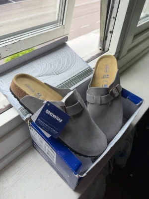 Gråa Birkenstock tofflor storlek 42 - Säljer ett par snygga gråa Birkenstock tofflor i mjuk mocka med justerbart spänne och klassisk korksula. Perfekta för dig som vill ha både stil och komfort hemma eller på språng. Levereras i originalkartong.