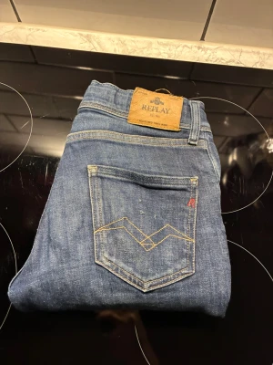 Blå jeans från Replay med slitningar - Säljer ett par klassiska blå jeans från Replay med snygga slitningar och detaljerade sömmar på bakfickan. Jeansen har fem fickor, knappgylf och är gjorda i denim. Perfekta för en avslappnad och trendig look. Ny pris 799