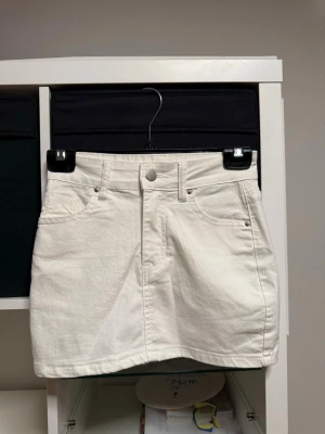 Vit kort jeanskjol med fickor - Snygg vit kort jeanskjol med klassisk femficksdesign och bälteshällor. Kjolen har knapp och dragkedja framtill samt raka sömmar. Hann tyvärr inte använda den innan jag växte ut den så har endast testat den.