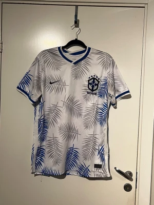 Vit Brasilien fotbollströja Nike - Snygg vit fotbollströja från Nike med Brasilien-emblem på bröstet. Tröjan har blå och grå palmbladsmönster över hela tyget samt blå detaljer vid krage och ärmslut. Perfekt för dig som vill sticka ut på planen eller heja på Brasilien.