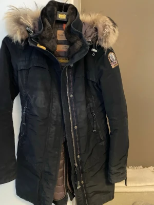 Parajumpers dunjacka  - Säljer en svart dunjacka från Parajumpers med avtagbar pälskant på huvan och flera praktiska fickor. Jackan har en lång, figurnära modell och detaljer som patch på ärmen och orange hängare i nacken. Perfekt för kalla vinterdagar. Orginal pris 6200. Perfekt för en kvinna under höst och vinter