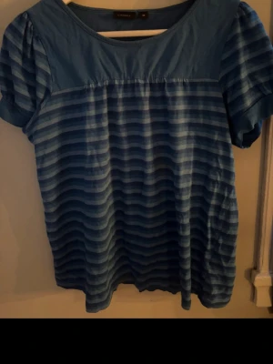 Blå randig topp med puffärm - Blå topp med horisontella ränder i olika blå nyanser. Modellen har rund halsringning och korta puffärmar. Tillverkad i mjukt material som känns skönt mot huden. Perfekt för dig som gillar en avslappnad men ändå snygg stil.