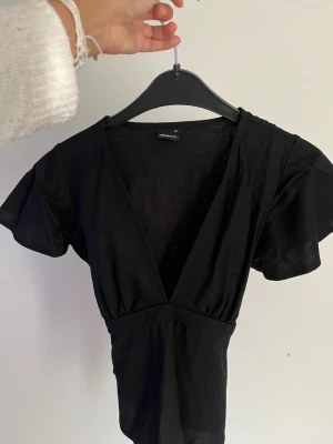 Svart omlottopp med knytband - Svart omlottopp med djup v-ringning och korta ärmar. Toppen har knytband i midjan som ger en snygg siluett och är tillverkad i ett mjukt, tunt material. Perfekt att styla med jeans 