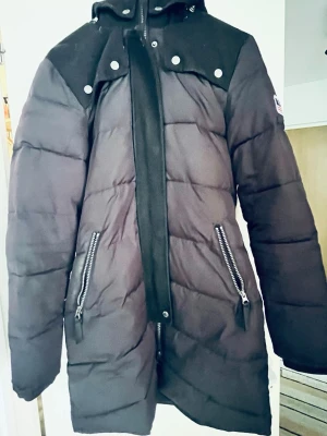 Svart dunjacka från Bondelid - Snygg svart dunjacka från Bondelid med huva och dragkedja framtill. Jackan har två snedställda fickor med dragkedja och dekorativa knappar vid axlarna. Perfekt för kalla vinterdagar och har en modern, längre passform.