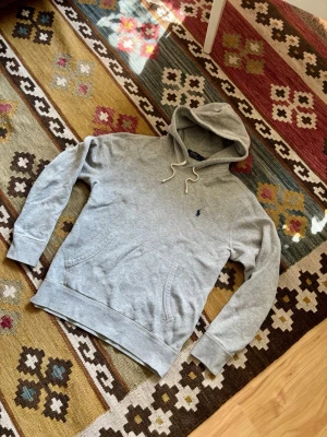 Grå hoodie från Polo Ralph Lauren - Säljer en klassisk grå hoodie från Polo Ralph Lauren med huva och dragsnören. Hoodien har en liten mörkblå logga broderad på bröstet och en stor magficka framtill. Tillverkad i mjukt sweatshirtmaterial och har ribbade muddar vid ärmar och nederkant. Nypriset ligger runt 2000kr!
