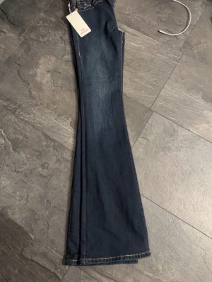 Mörkblå bootcut jeans med fickdetaljer - Snygga mörkblå bootcut jeans med markerade sömmar och stora bakfickor med dekorativa broderier och knappar. Jeansen har klassisk femficksdesign och en lätt utsvängd passform nertill. Perfekta för dig som gillar retrovibbar och streetstyle.
