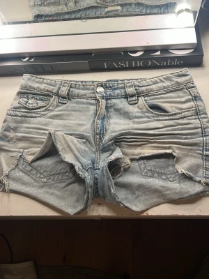 Ljusblå denimshorts med broderi - Säljer ett par ljusblå jeansshorts i denim med slitna detaljer och råa kanter. Shortsen har broderade bakfickor med paisleymönster och silverfärgade knappar. Klassisk femficksmodell och bälteshällor. Perfekta för sommaren!