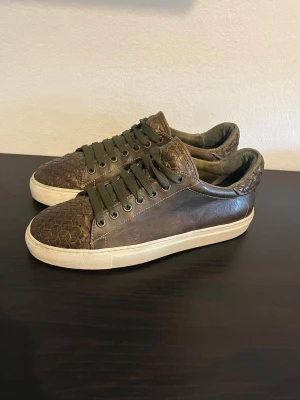 CC Shoes Sneakers – Kalvskinn | Mörkgrön/Olivgrön | Strl 40.5 (passar upp till 42) - CC SHOES SNEAKERS  Storlek: 40.5 — passar upp till 42 tack vare generös passform Färg: Mörkgrön/olivgrön med krokodilpräglad detalj vid hälkappan Material: Kalvskinn Sula: Vit gummisåla  Sneakers i äkta kalvskinn med en texturerad, reptilpräglad detalj vid hälkappan som ger dem ett unikt lyft. Mörkgrön ton som är sällsynt och lätt att kombinera med neutrala outfits — svarta byxor, beige chinos eller mörkt denim.  Skick: Bra skick. Normalt slitage på sulan, inga repor eller fläckar på ovandelen.