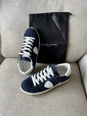 Philippe Model blåvita sneakers - Snygga sneakers från Philippe Model i marinblå mocka med vita detaljer och vita skosnören. Klassisk låg modell med vit sula och logotyp på sidan. Perfekta för dig som gillar stilrena och trendiga skor.